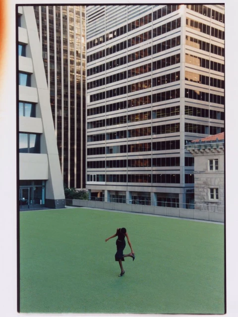 Woman in black dress poses on bright green rooftop lawn surrounded by tall office buildings in an urban setting on a sunny day.