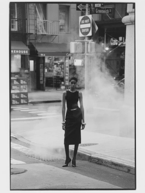 A slender Black woman wearing a dark dress and bracelets poses on a city street with steam rising from the road on a cloudy day.