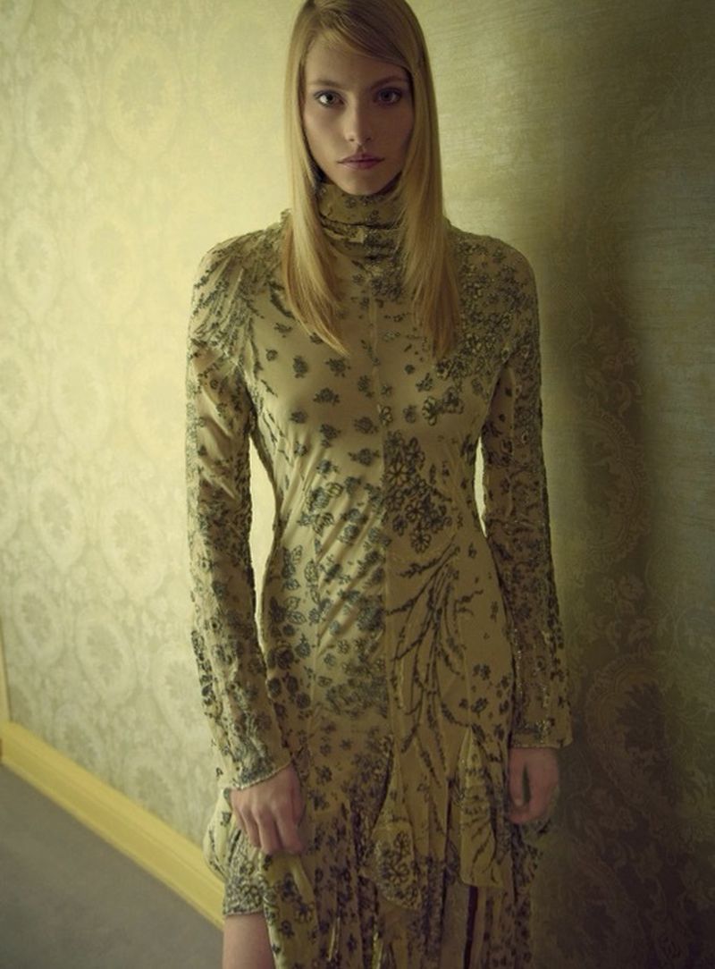Fashionable blonde woman in a patterned, long-sleeved turtleneck dress poses against a muted green wall with a decorative gold border.