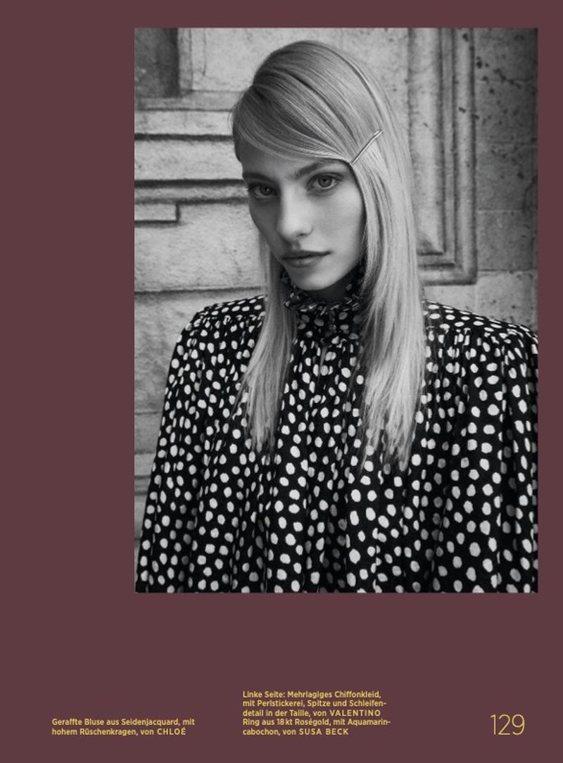 Portrait of an adult female model with blonde hair, wearing a black and white polka dot top in front of a neutral stone background.