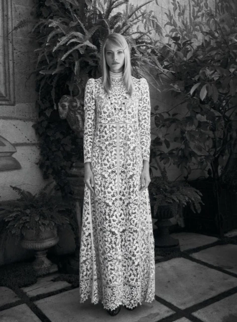 Full-length portrait of a woman with blonde hair wearing a long, patterned white gown, standing outdoors amid lush greenery in black and white.