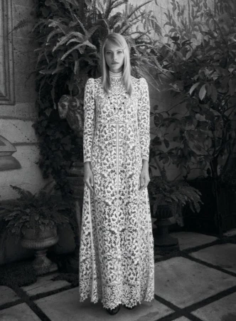 Full-length portrait of a woman with blonde hair wearing a long, patterned white gown, standing outdoors amid lush greenery in black and white.