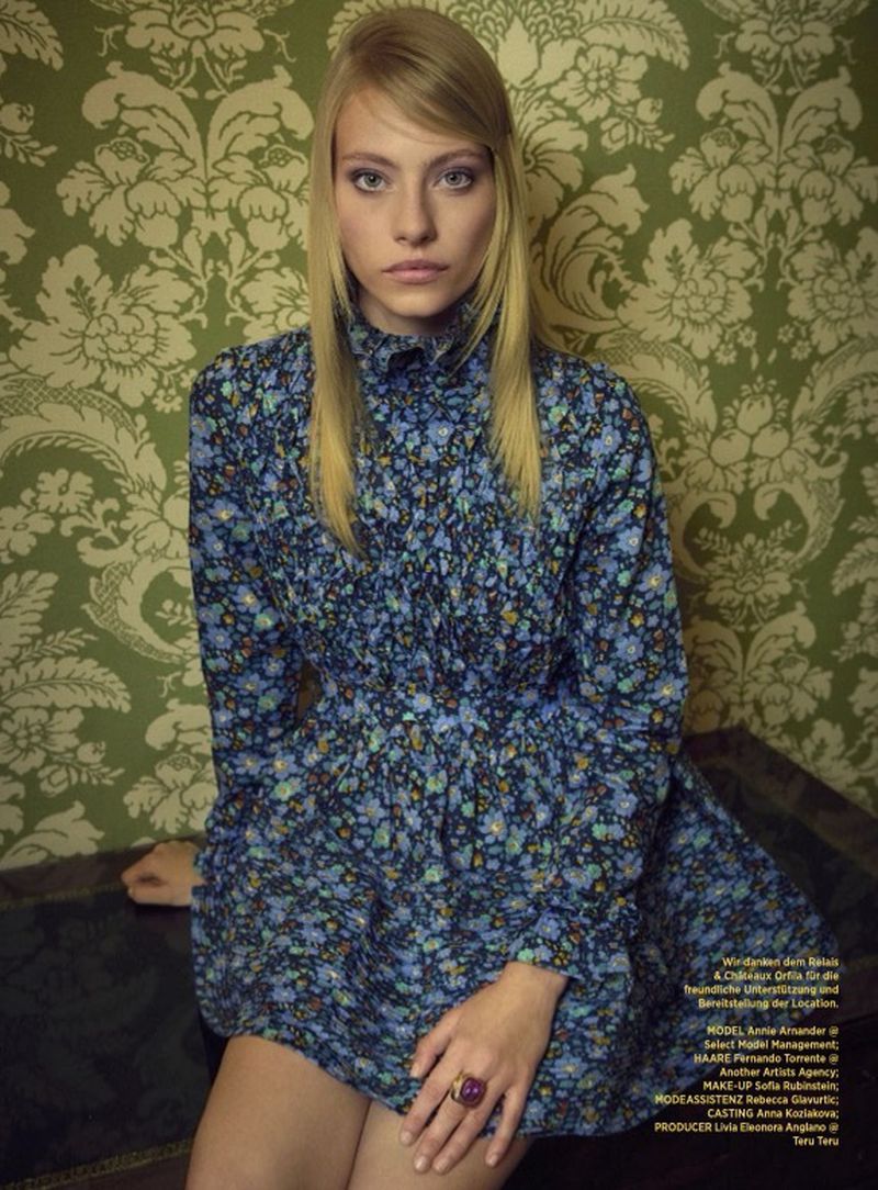 Model Annie Arnander in a long-sleeved blue floral mini dress sitting in front of green ornate damask wallpaper.