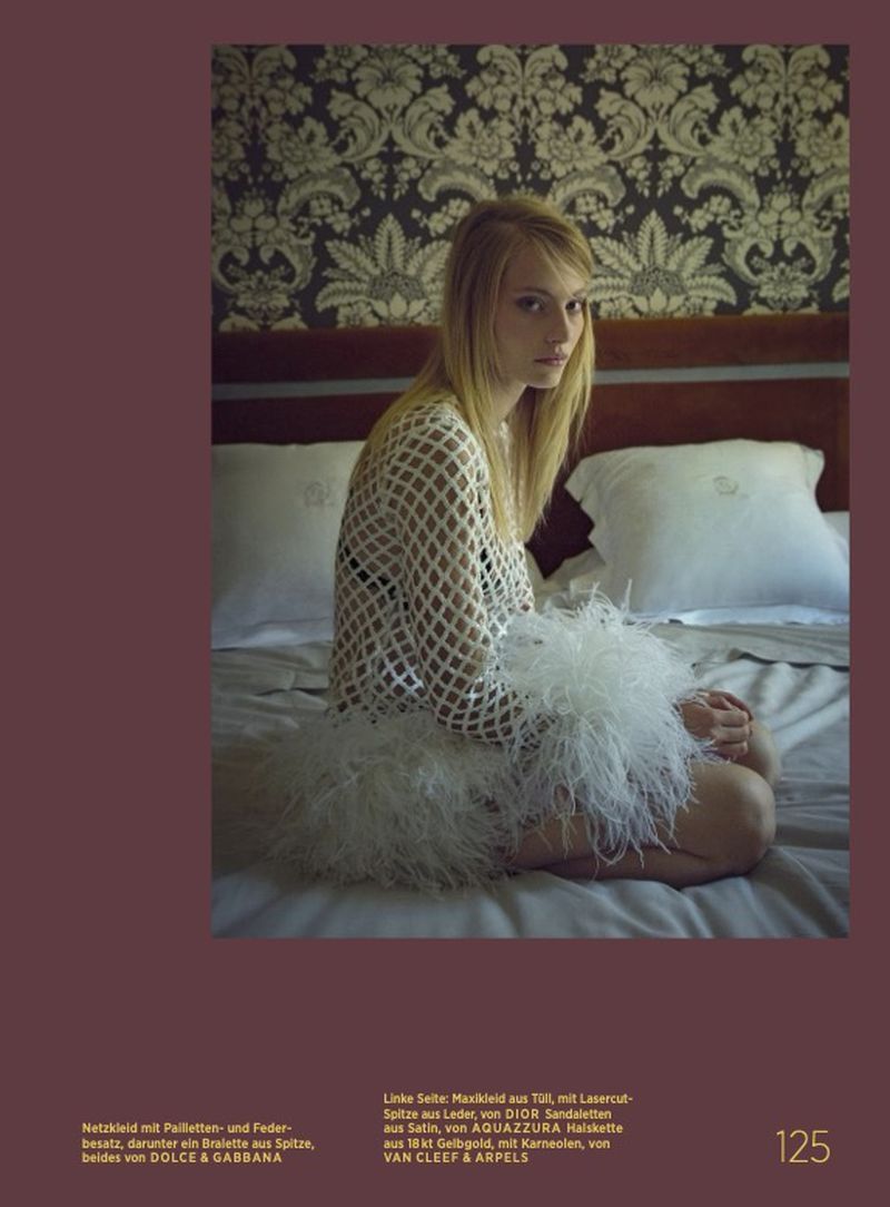 A fair-skinned woman with blonde hair sits on a bed in a net dress and feather skirt, with a patterned wall behind her.