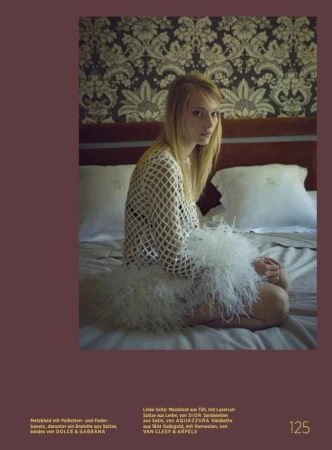 A fair-skinned woman with blonde hair sits on a bed in a net dress and feather skirt, with a patterned wall behind her.