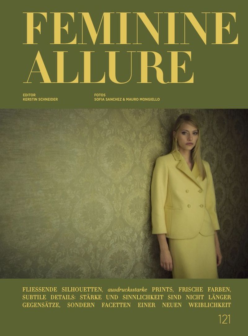 Editorial fashion cover featuring a blonde model in a yellow suit, set against a textured wall with olive green background and prominent typography.