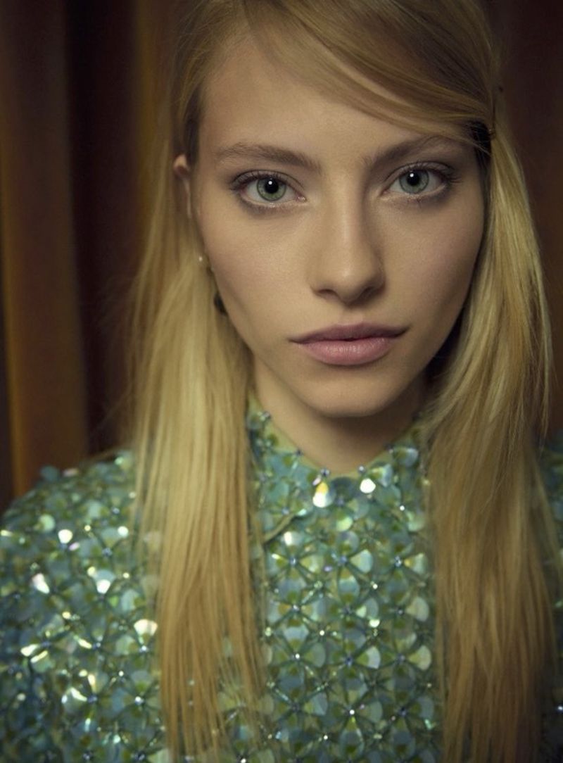 Close-up portrait of a blonde woman with light skin wearing a blue and green sequined shirt against a dark curtain background.
