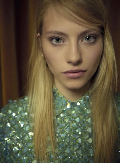 Close-up portrait of a blonde woman with light skin wearing a blue and green sequined shirt against a dark curtain background.