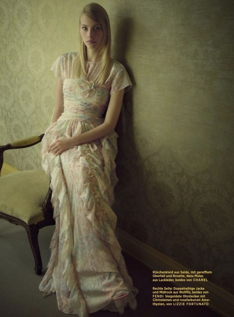 A slender blonde woman with long hair poses in a vintage-style floral gown in front of a textured green wall next to a vintage chair.