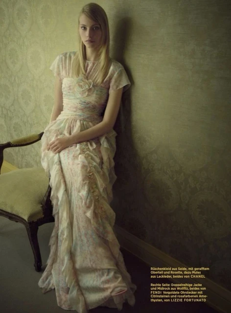 A slender blonde woman with long hair poses in a vintage-style floral gown in front of a textured green wall next to a vintage chair.