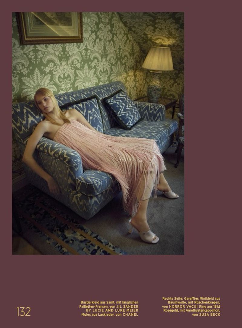 An adult female lies on a patterned couch in a fashionable pink dress with fringe. The room has patterned wallpaper and a lamp.