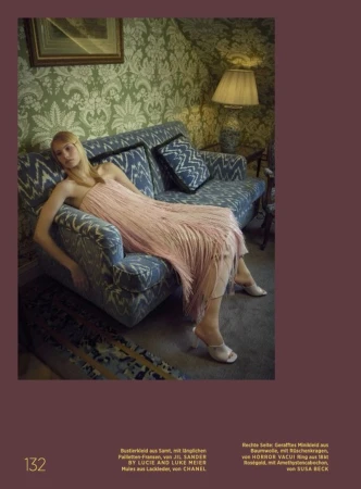 An adult female lies on a patterned couch in a fashionable pink dress with fringe. The room has patterned wallpaper and a lamp.