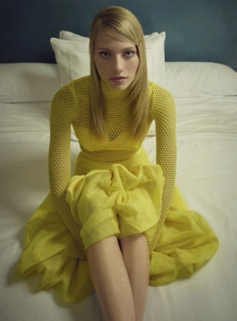 Model Annie Arnander wearing a bright textured yellow dress while seated on a bed with white pillows.