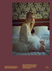 Model Annie Arnander seated on a bed in a white lattice mesh dress with feathery trim against patterned wallpaper.
