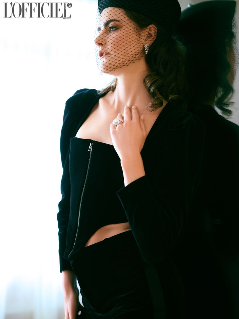 Elegant woman in black formalwear with sheer veil poses in a close-up portrait. High fashion editorial shot with neutral indoor backdrop.
