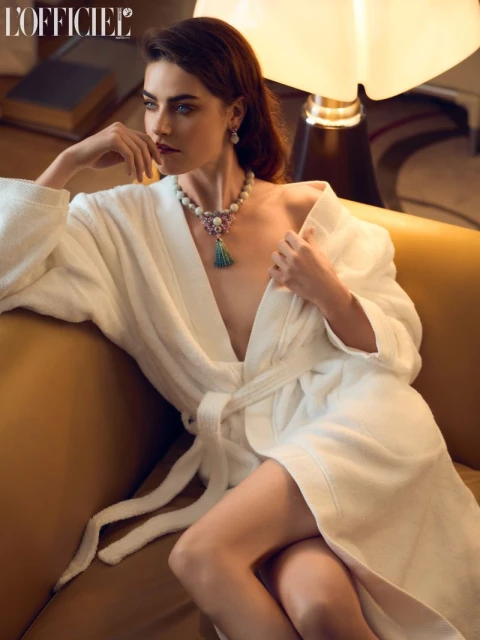 Elegant woman in a white robe rests on a gold sofa, adorned with a pearl necklace and earrings, in an indoor setting with soft lighting.