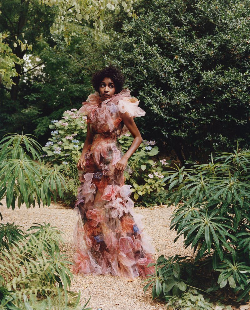 A woman in a ruffled gown poses in an overgrown garden. The dress features shades of orange, pink, and purple. Surrounded by green foliage.