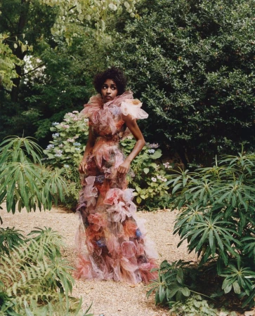 A woman in a ruffled gown poses in an overgrown garden. The dress features shades of orange, pink, and purple. Surrounded by green foliage.