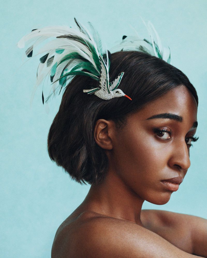 Close-up portrait of a woman with dark hair wearing a jeweled hummingbird and feather headpiece against a light blue background.