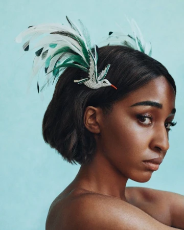 Close-up portrait of a woman with dark hair wearing a jeweled hummingbird and feather headpiece against a light blue background.