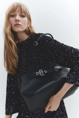Model with long blonde hair wearing a black sequin top and holding a black leather handbag against a white background