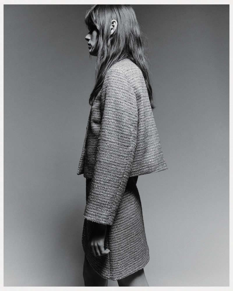 Monochrome side view of a slender adult woman in a tweed jacket and skirt, shot indoors against a gray backdrop in a fashion editorial.