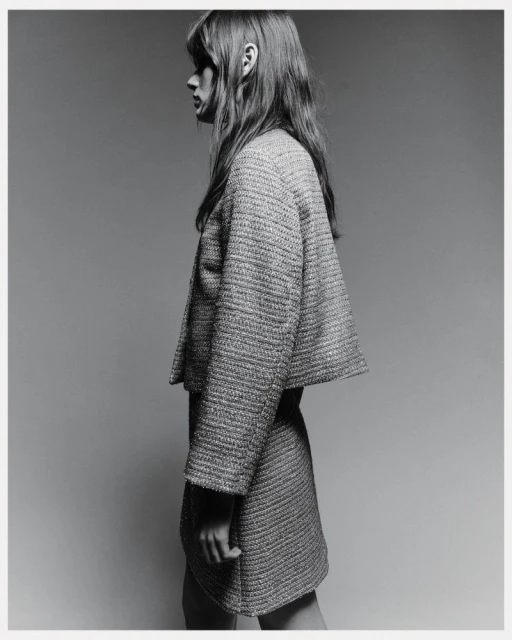 Monochrome side view of a slender adult woman in a tweed jacket and skirt, shot indoors against a gray backdrop in a fashion editorial.