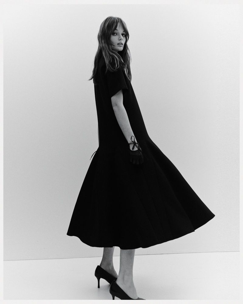 A slender woman poses in a black dress and heels against a neutral backdrop, her gloved hand subtly adorned with ribbon detail.