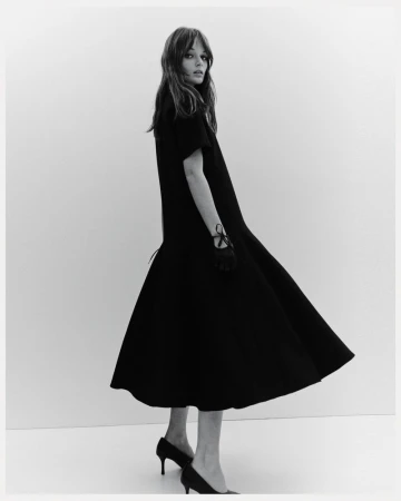 A slender woman poses in a black dress and heels against a neutral backdrop, her gloved hand subtly adorned with ribbon detail.