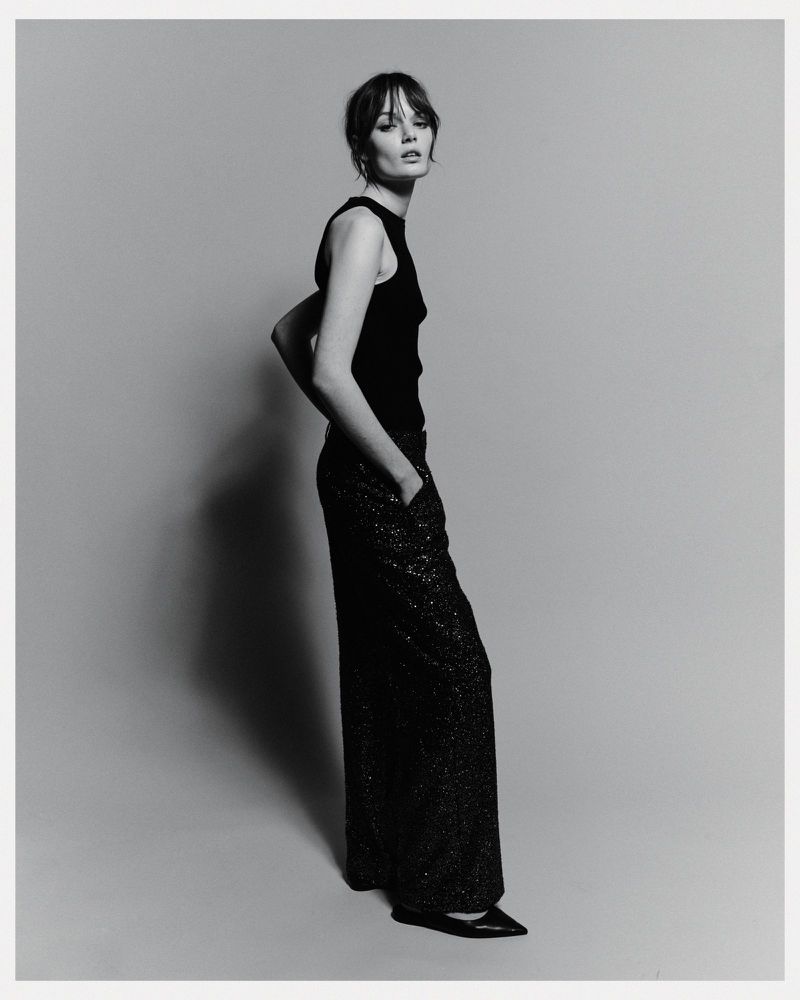 Model standing against a grey studio seamless backdrop wearing a black sleeveless top and dark sparkling trousers.