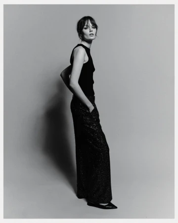 Model standing against a grey studio seamless backdrop wearing a black sleeveless top and dark sparkling trousers.