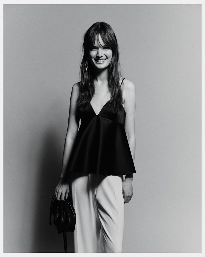 A woman with long brown hair smiles in a studio. She wears a black top, light pants, and holds a black purse in the monochrome image.