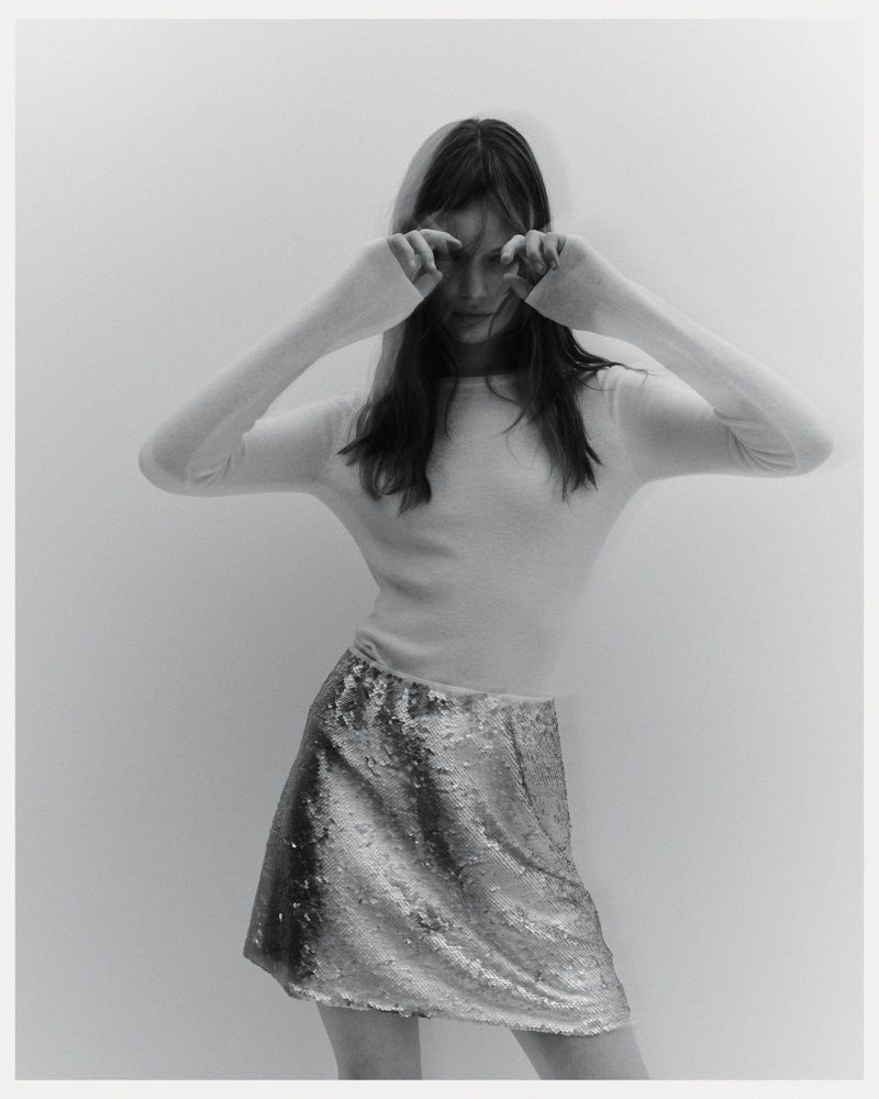 A young woman in a long-sleeved shirt and shimmering skirt poses against a neutral background, eyes closed with hands near her face.
