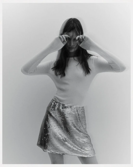 A young woman in a long-sleeved shirt and shimmering skirt poses against a neutral background, eyes closed with hands near her face.
