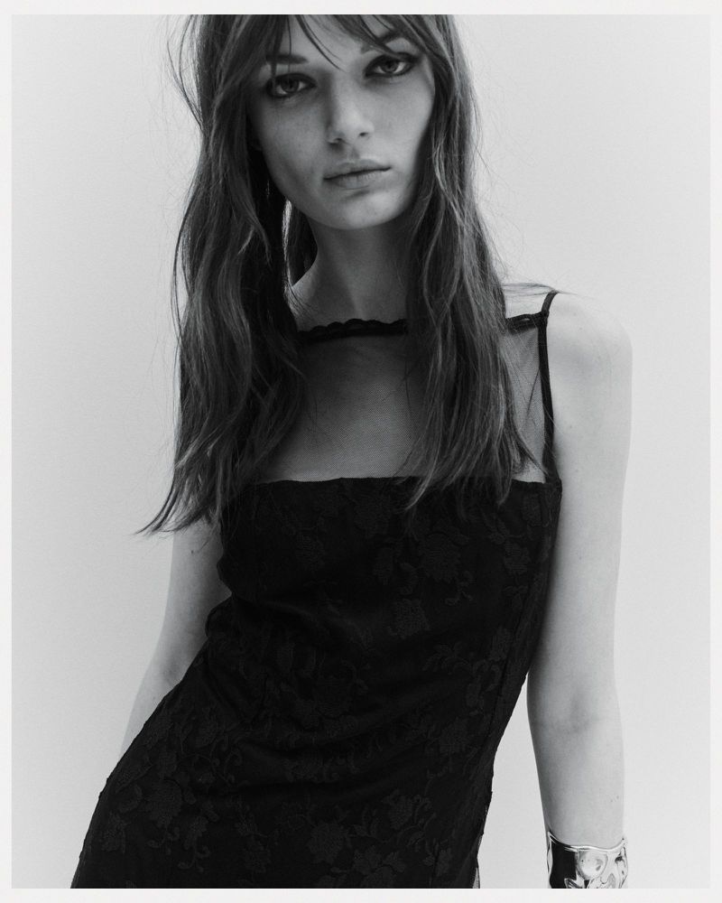 Monochrome portrait of a young woman with long wavy hair wearing a black lace dress and a metallic cuff bracelet, against a plain background.