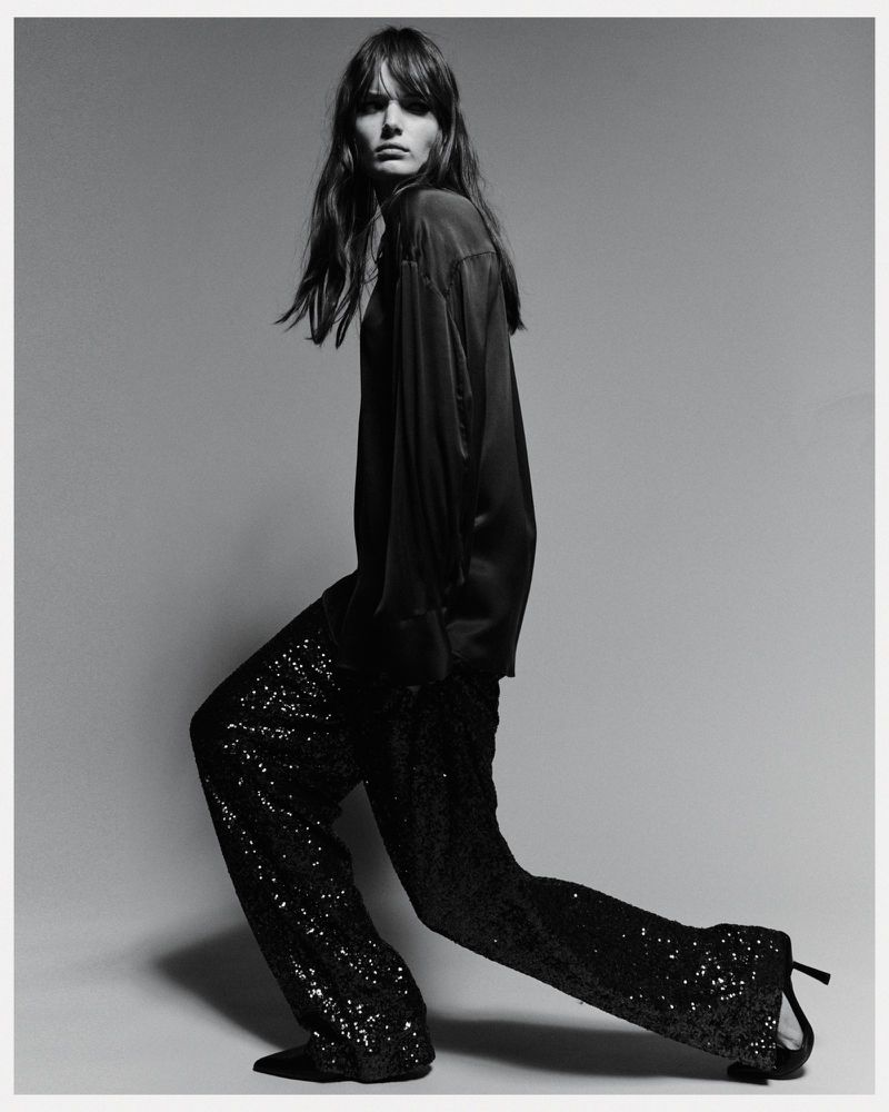 Black and white fashion shot of a woman in a dark top and sequined pants, posing in a studio with her body angled.