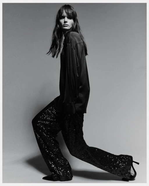 Black and white fashion shot of a woman in a dark top and sequined pants, posing in a studio with her body angled.