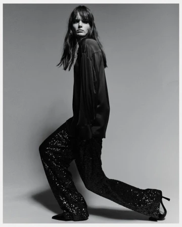 Black and white fashion shot of a woman in a dark top and sequined pants, posing in a studio with her body angled.