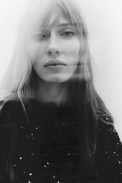 Black and white portrait of a blonde woman with bangs wearing a dark, sequined top. Soft, diffused lighting creates a hazy effect.