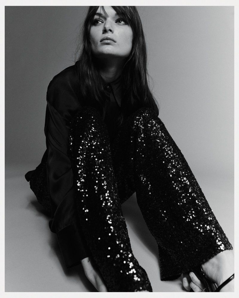 Portrait of a brunette adult female model wearing a black blouse and sequined pants in a studio with soft lighting and grey background.