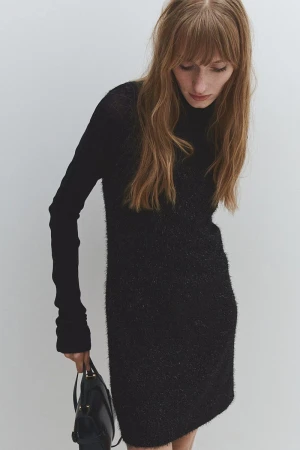 Model in black textured long-sleeve dress holding a black handbag against a light grey background