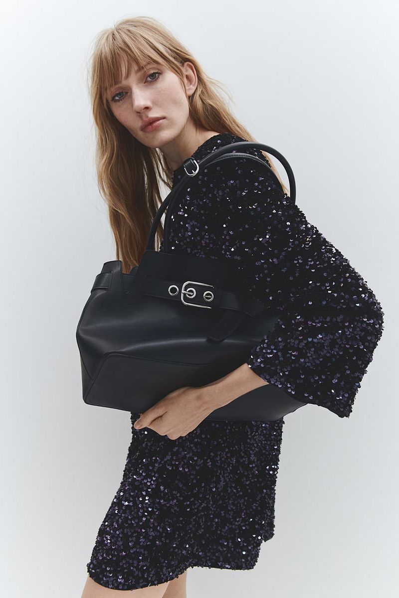 A fair-skinned woman with long blonde hair holds a black tote bag, wearing a short, long-sleeved, sparkly blue dress against a white backdrop.