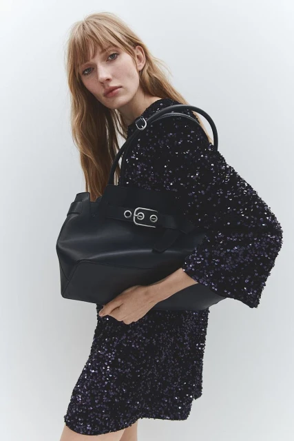 A fair-skinned woman with long blonde hair holds a black tote bag, wearing a short, long-sleeved, sparkly blue dress against a white backdrop.