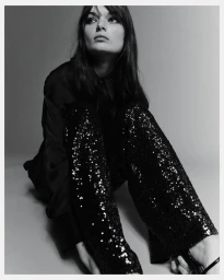 Model seated on the floor wearing black sequin trousers and a dark blouse against a plain grey studio background.