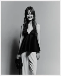 Smiling female model in black v-neck top and white trousers holding a small black handbag against a grey seamless studio backdrop
