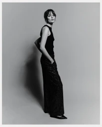 Model standing against a grey studio seamless backdrop wearing a black sleeveless top and dark sparkling trousers.