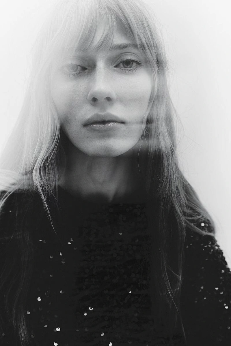 Close-up portrait of a model with blonde hair and bangs wearing a dark sequined top against a plain light background