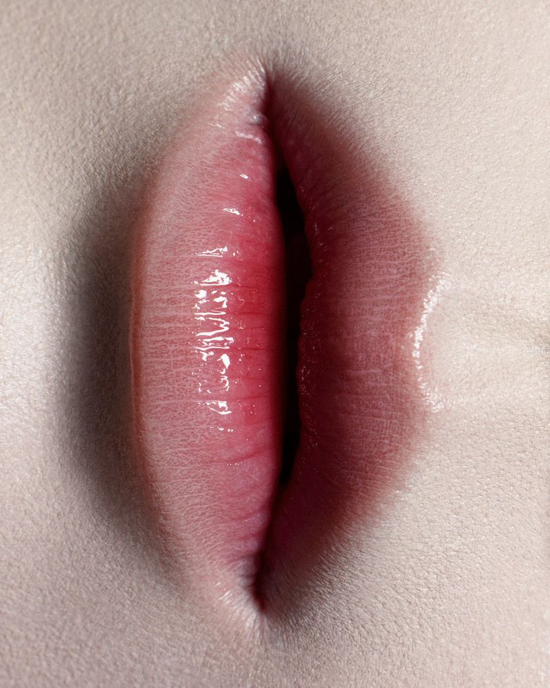 Close-up of human lips with a glossy, red-pink hue, highlighting texture and detail in a studio shot on a fair-skinned face.