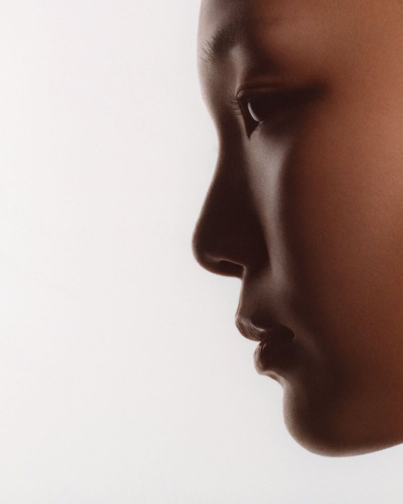 Extreme close-up profile portrait of a woman's face in soft shadow against a bright white studio backdrop.