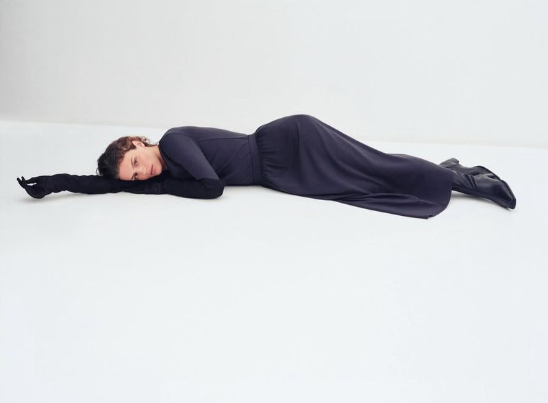 Woman in a long-sleeved black dress and gloves laying on a white studio floor, posing for a fashion shot with dramatic lighting.